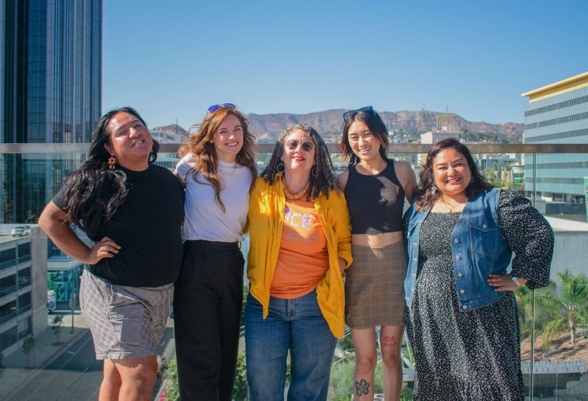 Photo of current LACE Team--two non-binary people and three women posed together for a group photo on a rooftop with a background view of the Santa Monica Mountains and Hollywood high rises. From left to right, a brown-skinned Latinx trans femme person, two fair-skinned Caucasian women, a non-binary East Asian person, and a light-skinned Latina all smile directly at the camera.
