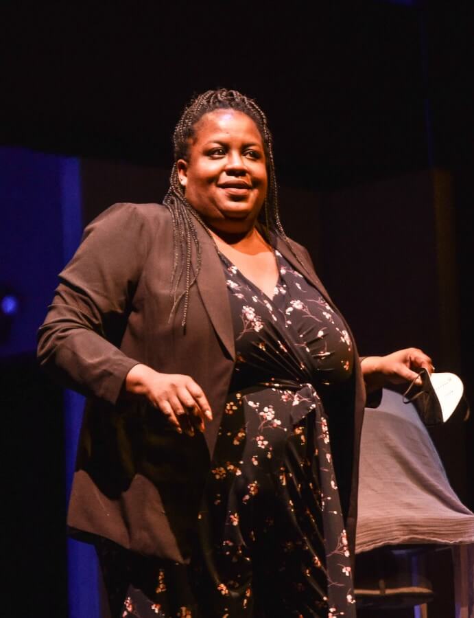 Lauren Turner Hines, a dark brown-skinned, full-figured Black woman, wearing braids a black flower-speckled jumper, and a black blazer jacket, smiles at the audience with joy while thanking them for attending the Opening Night of Draptemonia: A Negro Carol.
