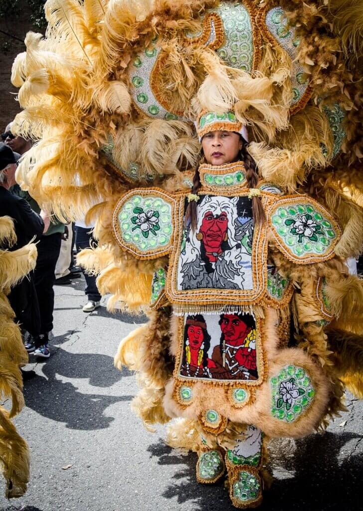 The image is a full-body portrait of a light-skinned African American woman wearing a gold-feathered Black Indian suit. Her headdress has gold feathers, green and white beads, and large green rhinestones. The stonework patches on her arms are of green and white magnolias, and the front patches show human figures in red, brown, white, and black beads. She is standing on the street surrounded by a crowd of adorning fans on a sunny day.