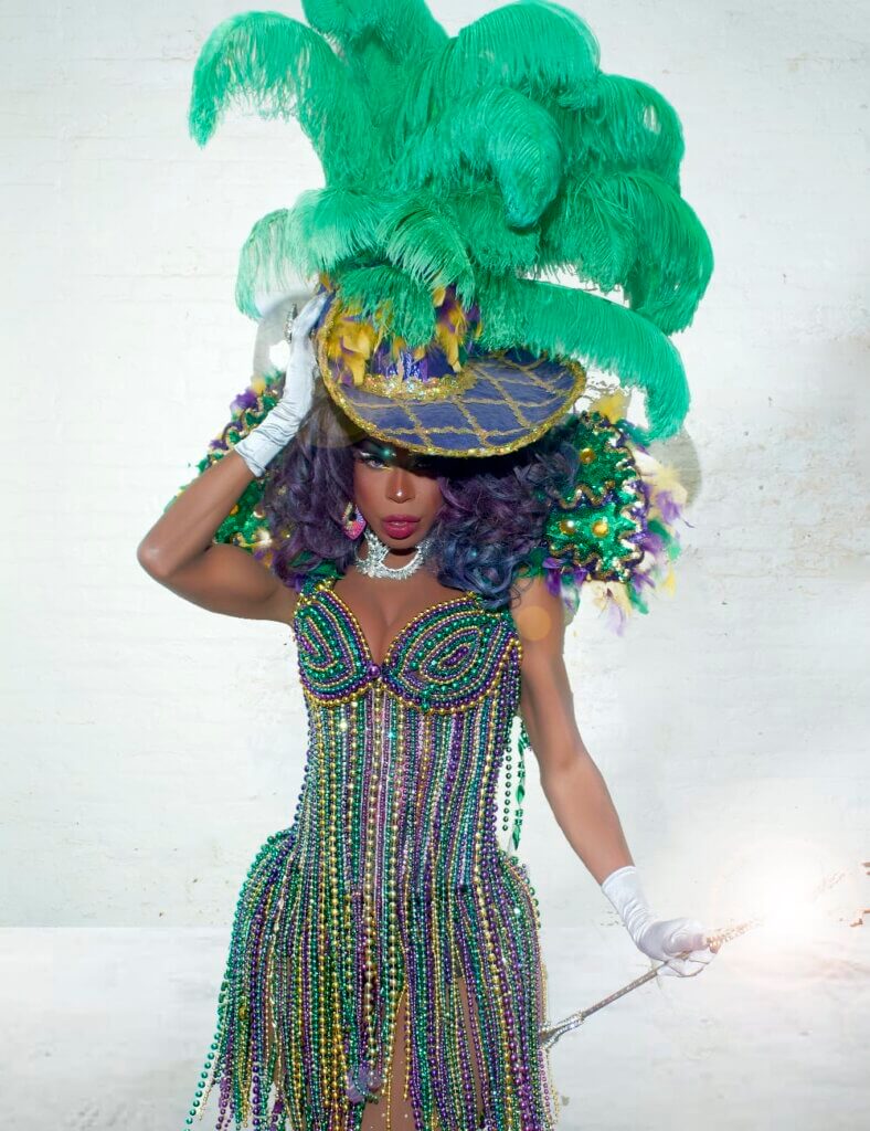 An image of a medium-skinned African American woman wearing an extravagant Mardi Gras-inspired outfit. The ensemble features a beaded corset dress adorned with purple, green, and gold beads, forming a fringe-like pattern, white opera gloves, and a purple hat with gold trim and large ostrich feathers. The background is a plain white wall, and she holds a baton in one hand.