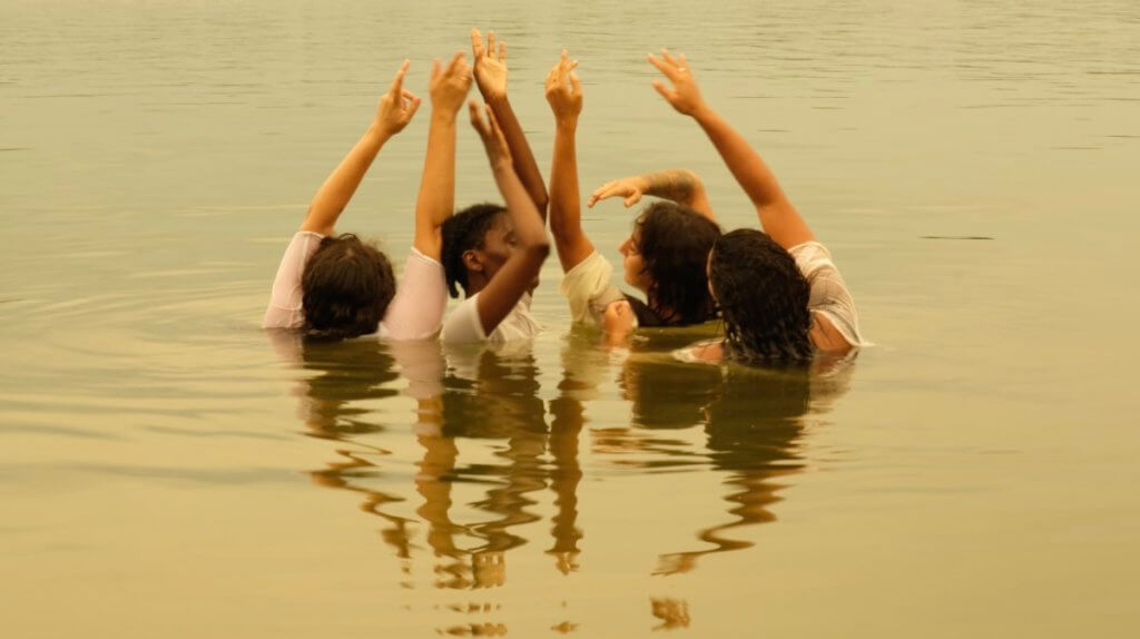 The image is sepia toned. Four dancers are shoulder deep in water with Black and brown arms reaching towards one another, soft hands, and gentle meditative gaze. Their shirts are white, and their reflection is mirrored upside down, in wavy ways on the water.
