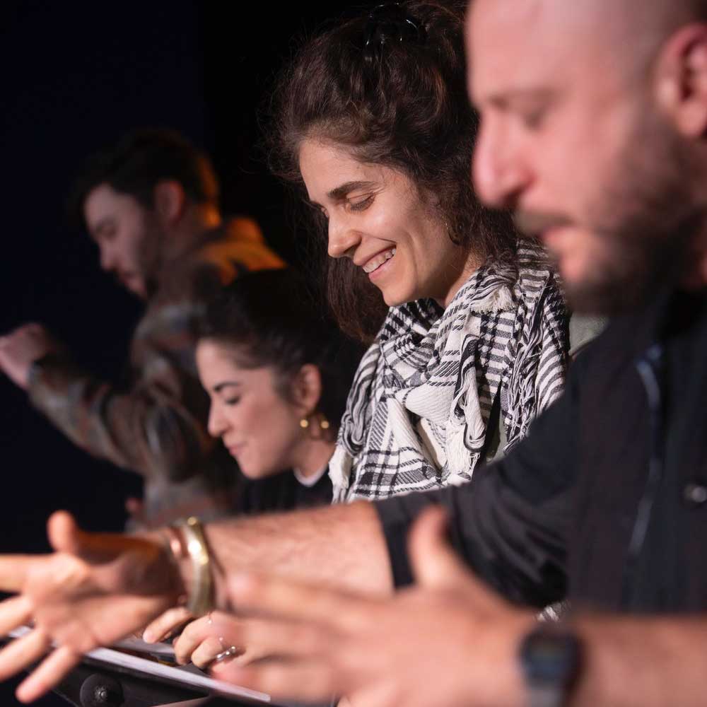 A close-up from the side of four actors standing in a line and reading from manuscripts as they perform. The background is dark and they're brightly lit as if on a stage. In the extreme foreground is the out-of-focus silhouette of the first actor, a man, who is gesturing with both hands. In the middle-ground in sharp focus is a female actor wearing a black and white plaid scarf. She is smiling as she looks down at her manuscript. Behind her in the background, both out-of-focus, is another female actor looking down and a male actor with his hand raised as he performs.