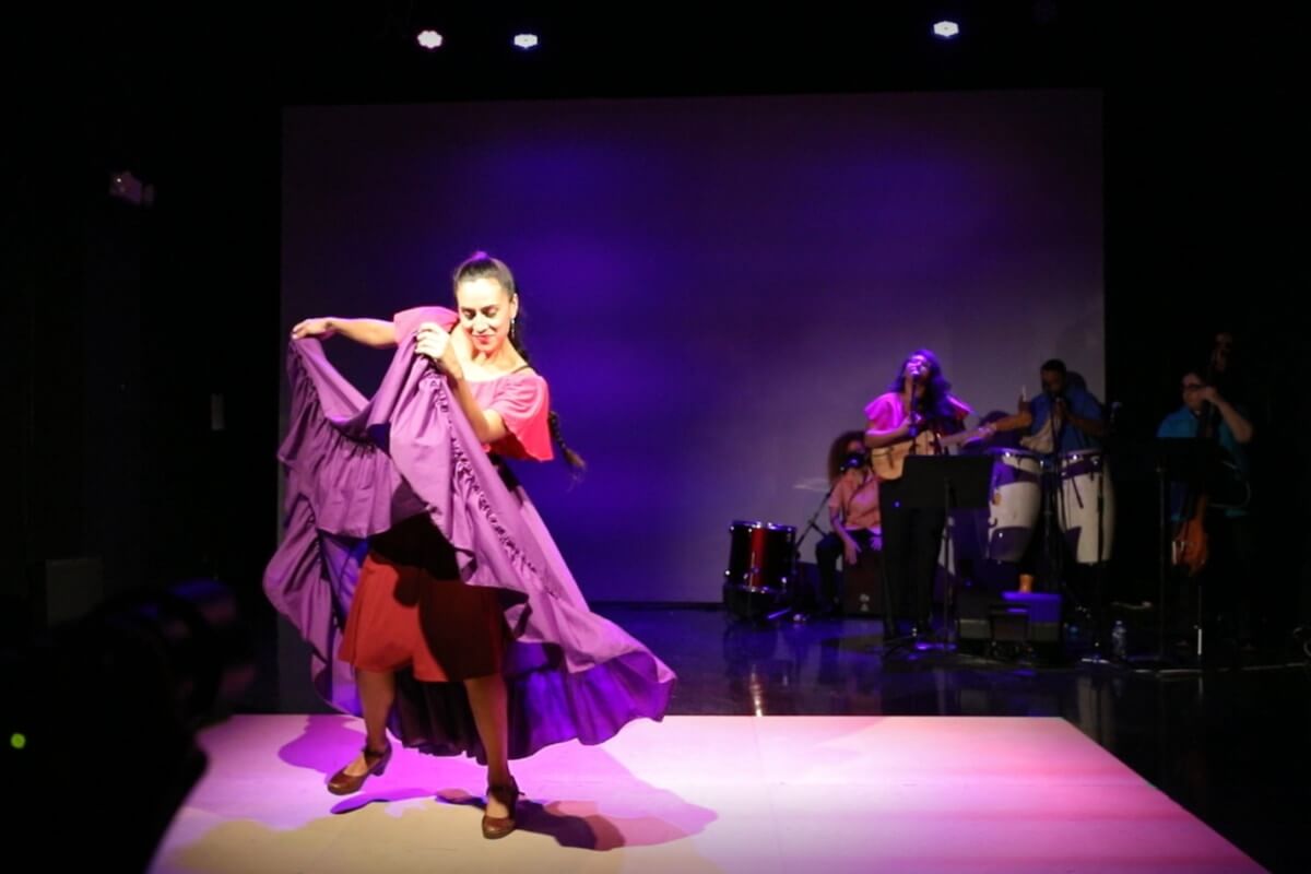 The photo depicts a Latinx woman mid-movement dancing in the foreground. She is on a wooden platform with her hands holding her purple skirt as she performs. A purple spotlight is on her. In the background, there are four musicians – three percussionists and one guitarist/vocalist.
