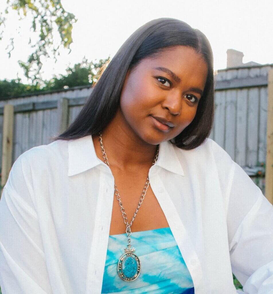 The image shows an Indigenous woman with long, black, straight hair. She is looking at the camera with her head tilted to the side. She wears a white button-down shirt, an aqua tank underneath, and a silver and turquoise jewelry necklace.