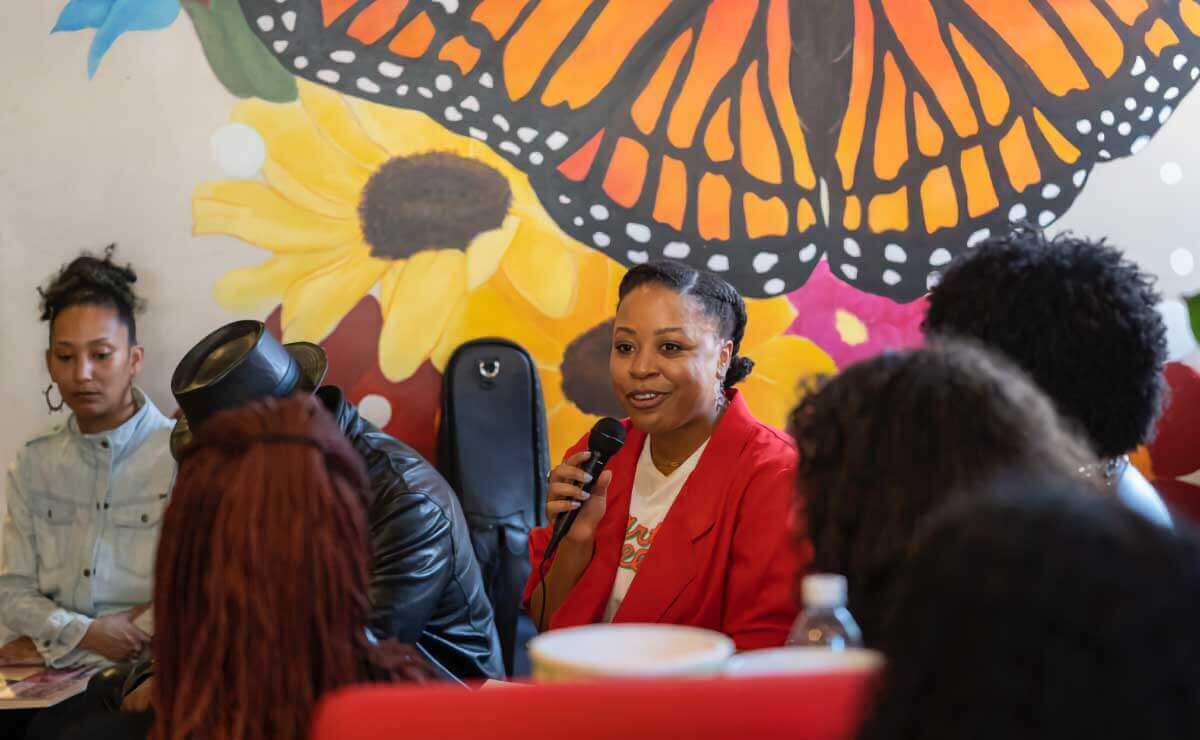 A small group of 7 people sit together. Most are in profile from behind. They sit against a colorful wall with a large butterfly painting. At the center, facing the camera, is a Black woman smiling, wearing a red blazer and white t-shirt.