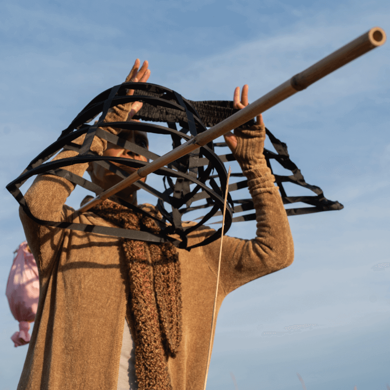 A person stands outdoors under a blue sky at midday, covering their head and shoulders in what appears to be a crinoline dress cage made of black, lattice-like material. They are dressed in an adobe brown sweater and a textured darker brown scarf, with a bindle stick over one shoulder and a knotted pink bindle bag tied to the end. The lattice object partially obscures their face, creating a dynamic and abstract composition. The scene is lit by warm sunlight, highlighting the textures and materials.