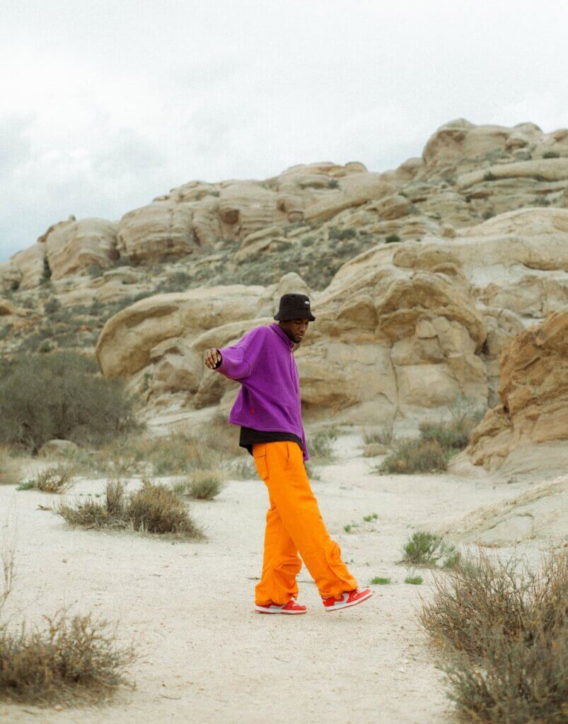 This image shows a dark-skinned man in mid-motion in the desert. His right arm is raised to shoulder level, and his right foot is slightly raised off the ground. He wears orange pants, a purple sweatshirt, and a black hat.