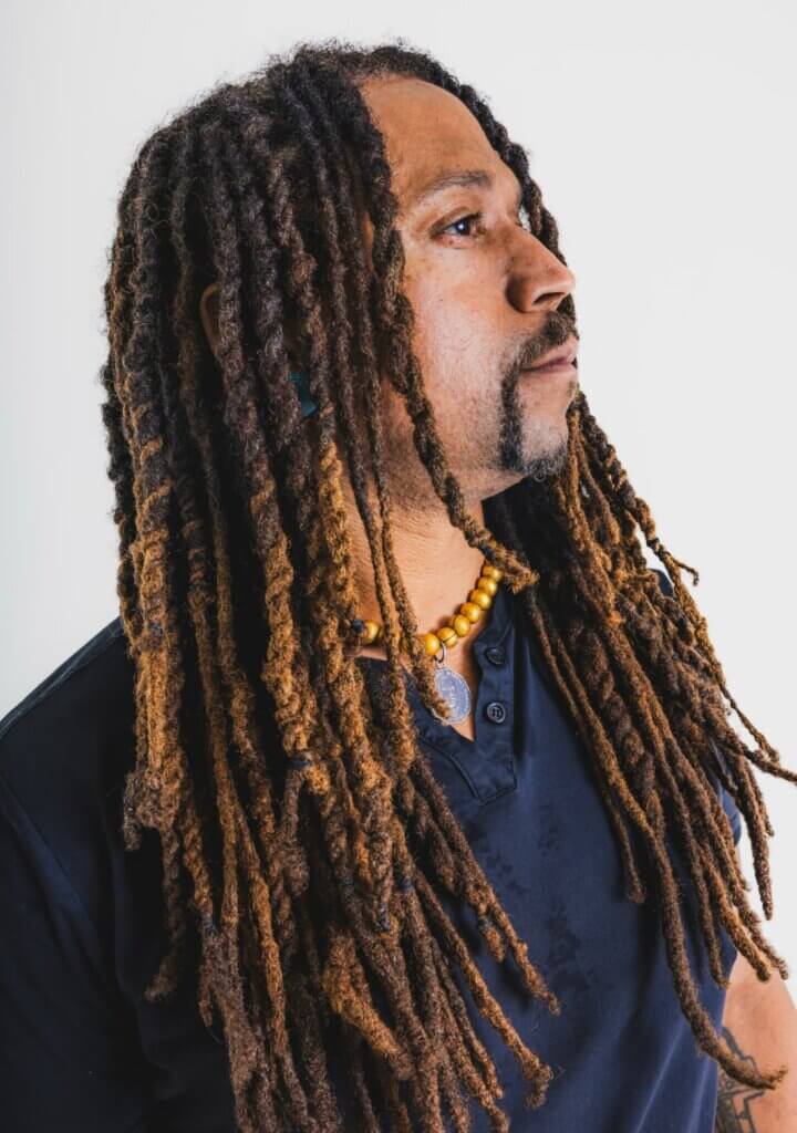 A light brown-skinned African American man standing with his head tilted to the sky. He has long, brown, twisted locs and a gold-beaded necklace around his neck.