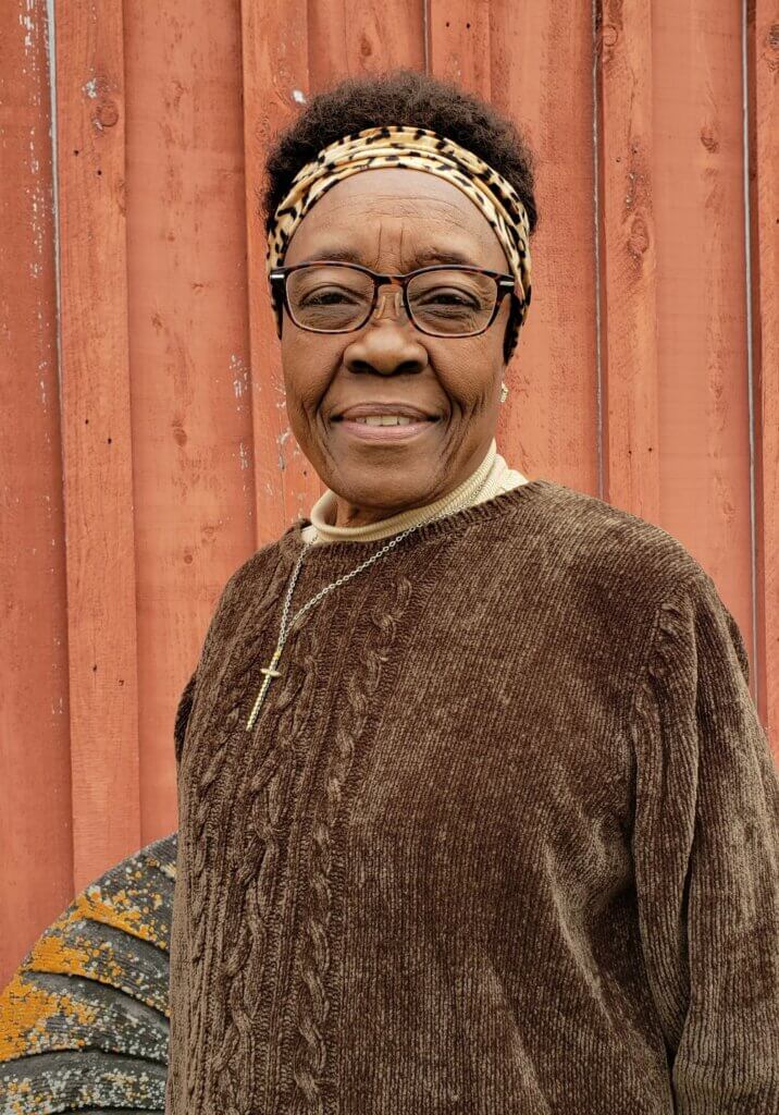 The image is of a medium brown-skinned African American woman. She is wearing a brown velour sweater, tan turtleneck underneath, black-rimmed eyeglasses, and a leopard print headband. The woman is standing outside in front of an old red barn.