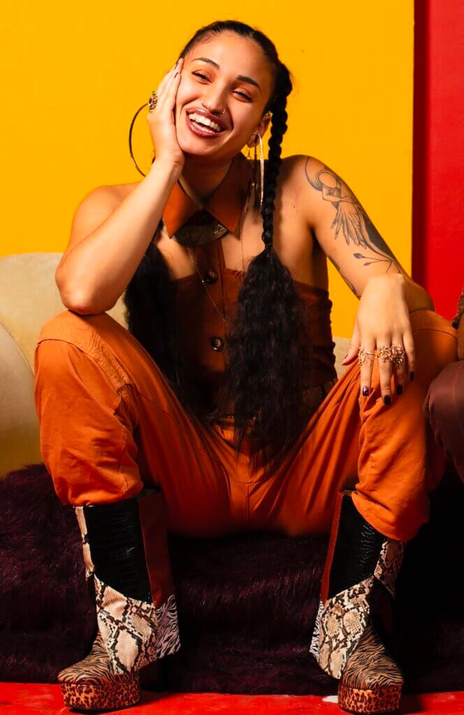 The image is of a tan-skinned person smiling while sitting on a couch with elbows resting on their knees and chin resting in their right hand. Their hair is in two braids. They wear a brown, collared button-up shirt vest, orange pants, animal print boots, large hoop earrings, and rings on their fingers.