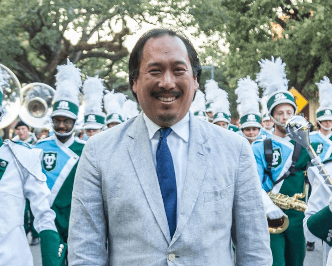 Mendel Lee dressed in a white suit and blue tie, standing in front of a marching band.