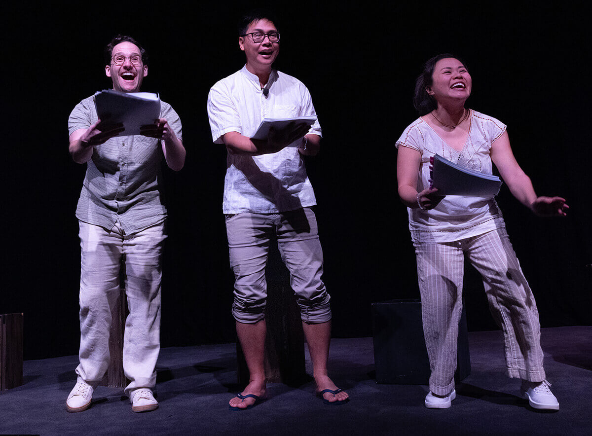 A trio of actors onstage with scripts in hand: a white man with glasses, a medium light-skinned Filipino man with glasses, and a petite medium-skinned Filipino woman stare up to the sky, wide, joyful smiles on their faces. They wear pants and shirts in whites, tans, and light browns.