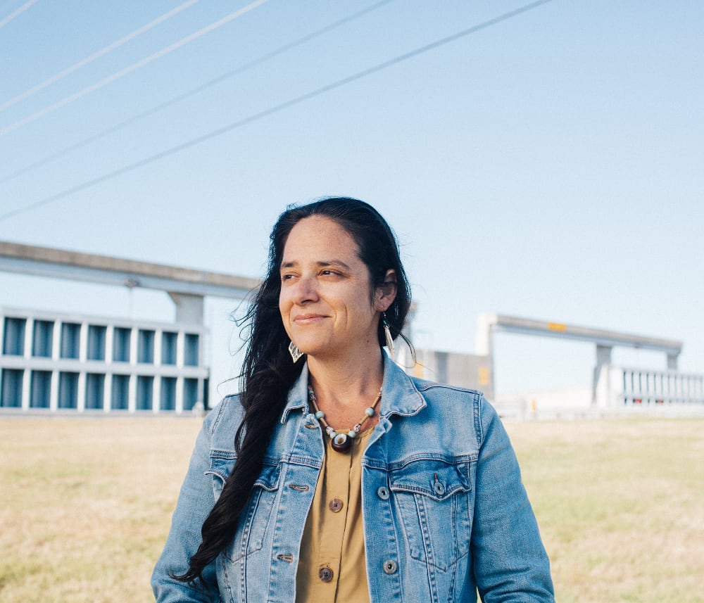 An image of an Indigenous woman in a jean jacket standing before a concrete flood levee wall. She has long brown hair, a mustard-colored blouse, and a seed pod necklace.