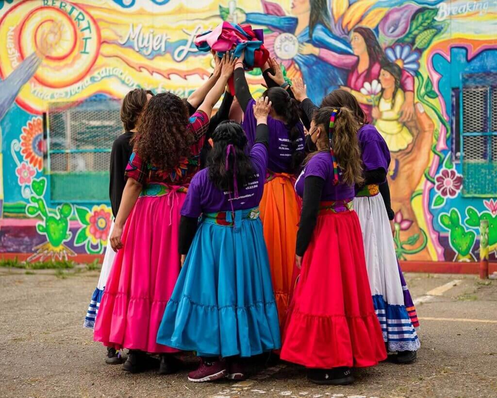 Eight women and nonbinary people are wearing brightly colored long skirts with ruffles in red, white, magenta, teal, orange. They are in a tight circle, outdoors, with their backs to the audience, holding up a ball of colorful ribbons. Behind them is a colorful mural with flowers, a sun, and the words 'Rebirth' and 'Mujer' (woman).