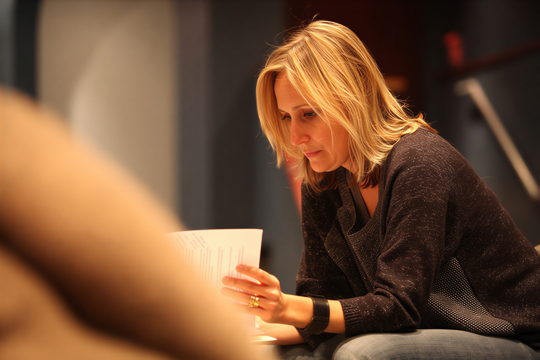 A blonde woman sits leaning forward while reading a printed document, focused on the pages in her hands. Warm indoor lighting and a softly blurred foreground create the feeling of a quiet discussion, rehearsal, or speaking event.