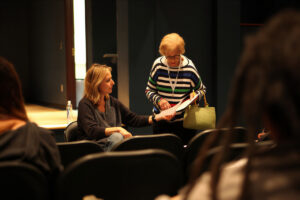 A seated woman hands a document to an older woman standing beside her in a small auditorium while attendees watch from nearby rows. The two appear to be discussing or reviewing paperwork during a meeting, workshop, or rehearsal in a softly lit event space.