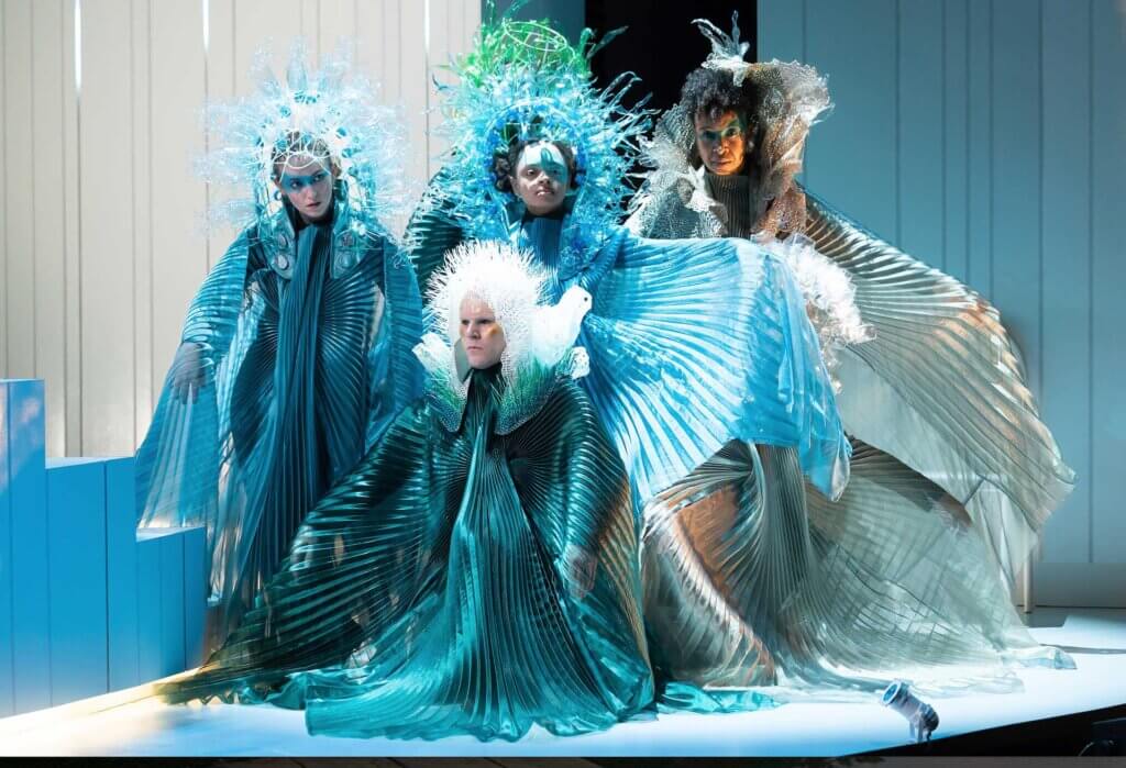Six actors - a mix of African American and white bodies, wearing flowing robes and multi-colored headdresses made from recycled plastic, resembling ocean corals and sea creatures.