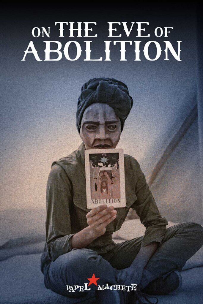 A brown-skinned, femme, masked person sits crosslegged in a tent with the words 'On the Eve of Abolition' above her and 'Papel Machete' below her. She holds up a large tarot card with the word 'Abolition' on it and stares directly into the camera. She is wearing a green jumpsuit and a black head wrap.
