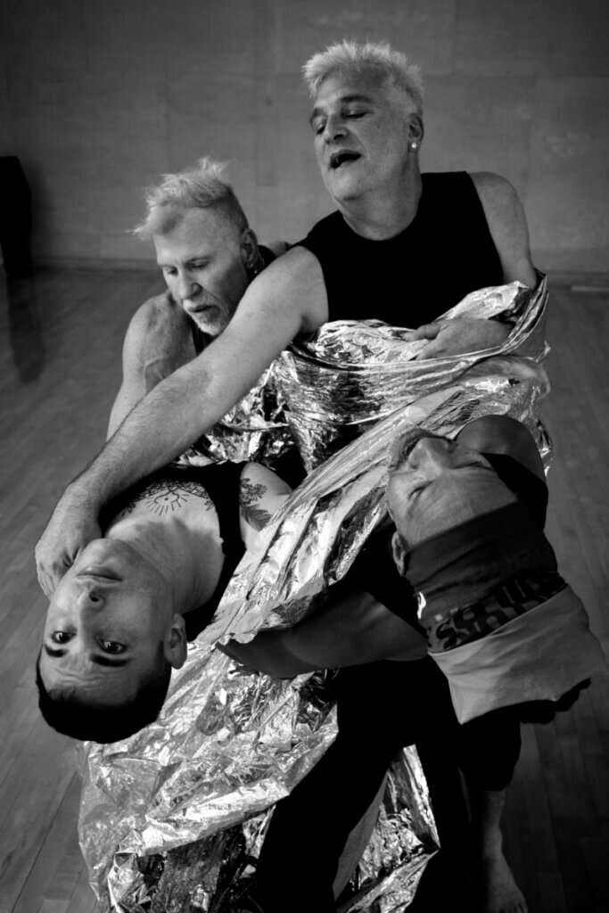 A black and white image capturing a moment of four dancers in a dramatic counter-balance, bound together by silver reflective fabric. Pioneer Winter, 36 year-old white genderqueer with tattoos on their chest and clavicles, is arched back next to Clarence Brooks, a 63 year-old medium-dark skinned Black genderqueer man with dreadlocks bound up like a crown atop their head. Both Pioneer and Clarence are supported by Cuban-American 59 year-old, silver haired Octavio Campos and 65 year-old white, fair skinned Frank Campisano with short, silver hair.