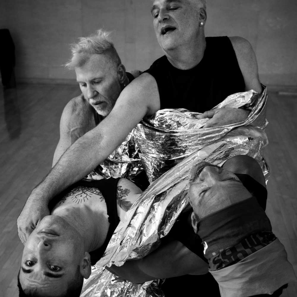 A black and white photo of four male dancers intertwined and bound together by a long bolt of what appears to be mylar or other similarly reflective silver fabric.