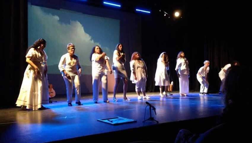 Nine Caribbean women performers vocalizing on stage. Standing in front of a screen projecting clouds in the sky. They are wearing white costumes. Minimal props. Dark blue light design. Musical instruments: guitar, percussion. Sound equipment on the floor. Part of the audience is visible from the left side.