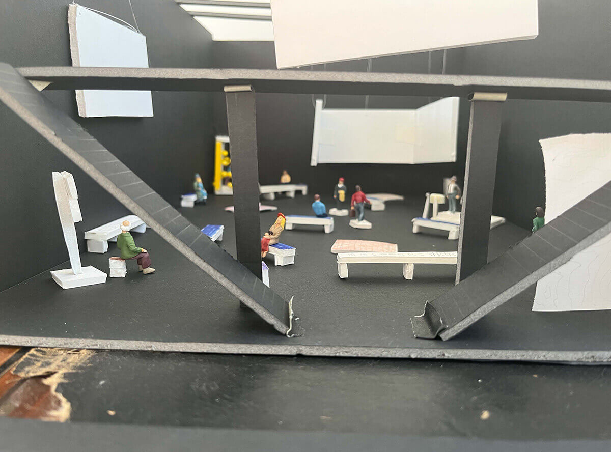 A quarter-inch scale model of a black box theater is filled with benches, screens, and models of people (some sitting and some standing). In the back corner there is something very bright and yellow, although it is blurry and hard to tell what it is.