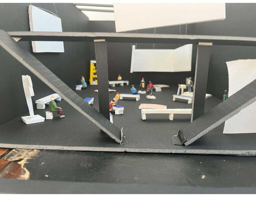 A quarter-inch scale model of a black box theater filled with benches, screens, and models of people (some sitting and some stranding), and in the back corner there is something very bright and yellow, although it is hard to tell what it is.