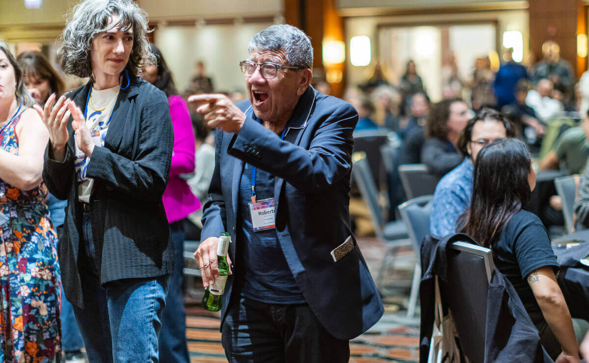 An older Latino man with short salt-and-pepper hair and glasses, and wearing a blue sports coat and blue shirt, stands In the center of a large convention hall and points happily to something out of frame. He is holding a bottle of beer in his right hand and pointing with this left hand. People standing nearby are applauding as they look at him.