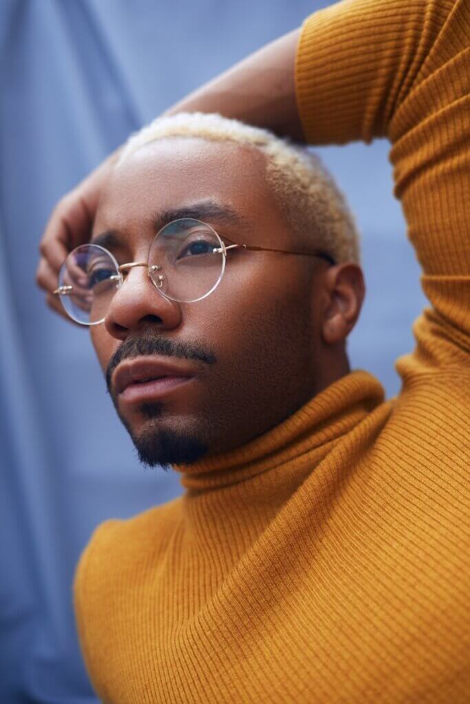 Roderick George posing wearing a fall yellow turtleneck looking in the distance while wearing glasses. The photo is a close up of his face and upper body.