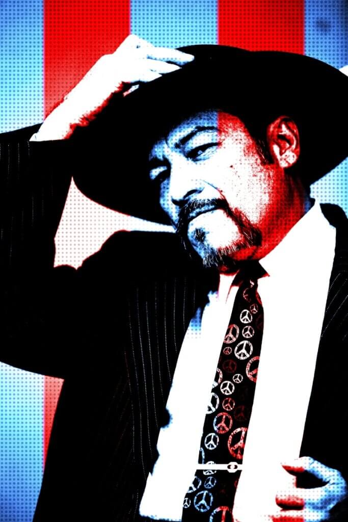 A medium skinned Chicano man dressed in a pin-striped suit, with a white shirt and a multi-colored tie that features round peace symbols. His facial features include a moustache that connects to a goatee. His left hand holds his left lapel, as his right hand reaches for his black, wide-brimmed pork pie Tando (hat), as if about to remove it from his head.