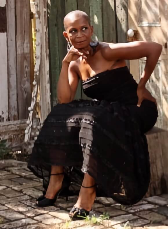Greer E. Mendy, a Black woman with medium-tone skin, sits on a wooden bench on a stone patio. She is wearing a black, strapless gown with black pumps. Leaning forward, her left hand is on her hip and her right elbow is on her knee, with her hand on her chin as she looks back toward her left.