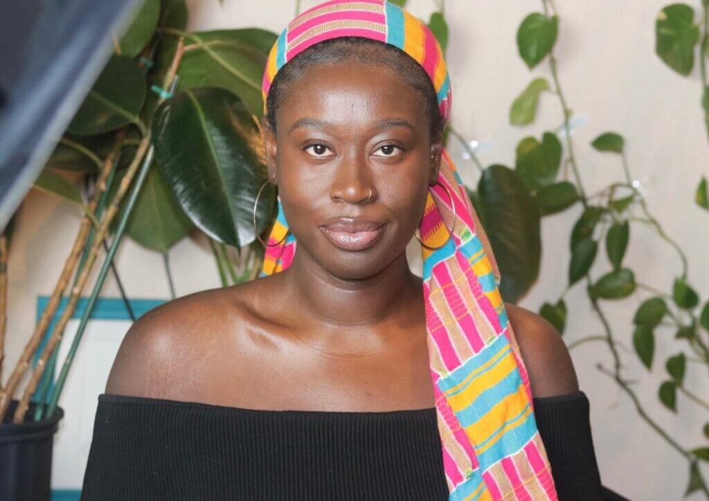 Nana Kumi, a Black woman with dark brown skin, looks directly into the camera. She is sitting proudly in her childhood home in Mississippi. Behind her is a light-colored wall covered in plant vines. She wears a pink, yellow, and blue kente cloth headscarf, silver hoop earrings, and an off-the-shoulder black top.	