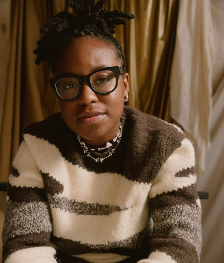 The photo show Sydney A. Foster, a Black woman with brown skin, dark brown locs, and wearing large, black glasses sitting for a portrait against a cream and gold curtain backdrop. She is wearing an abstract brown and cream sweater with brown pants.