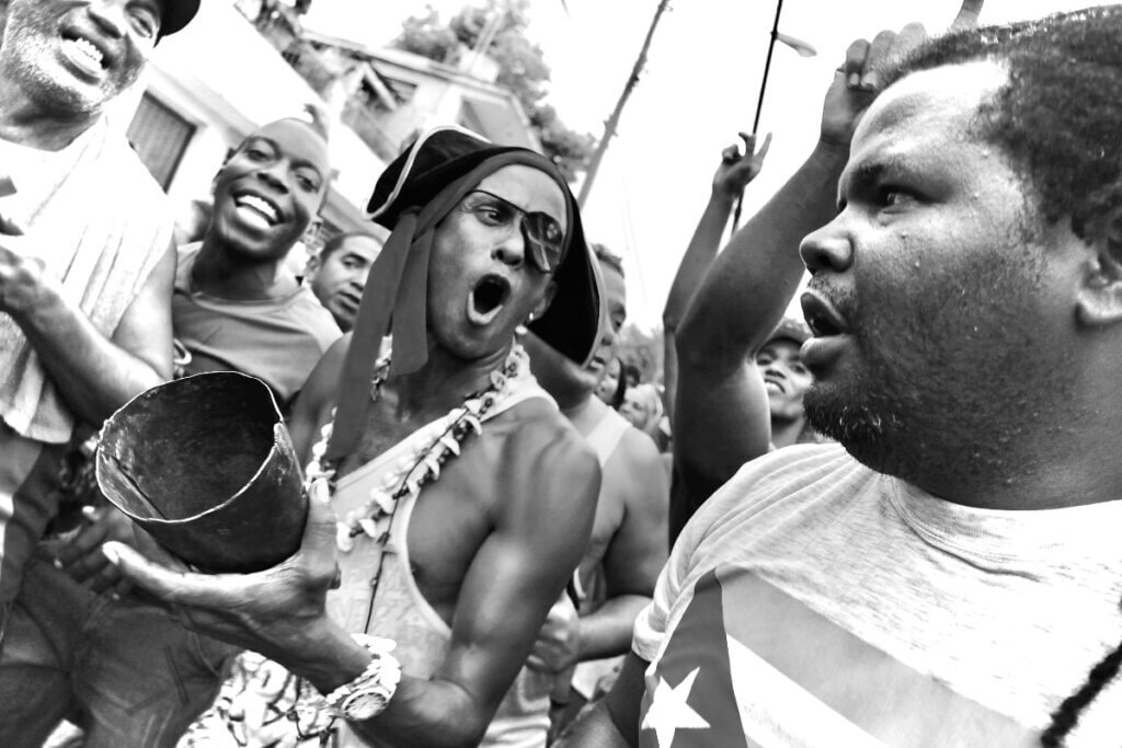The black and white photo depicts a man with dark skin, an eyepatch, and a pirate hat playing a cowbell. He and several onlookers look to their left with surprise and delight at the camera and a man with dark skin wearing a T-shirt with the Cuban flag.