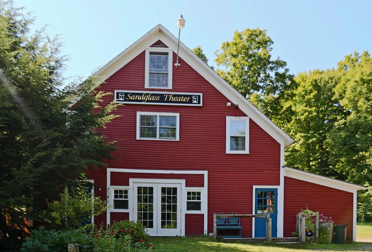 The photo depicts a red barn with white trim. A sign with gold lettering on a black background in the upper center of the building's facade reads “Sandglass Theater.” The theater has a blue door with two light blue seats next to it. Lush green plants and trees and several red and pink flowers surround the building on a sunny day with a wide blue sky in the background.