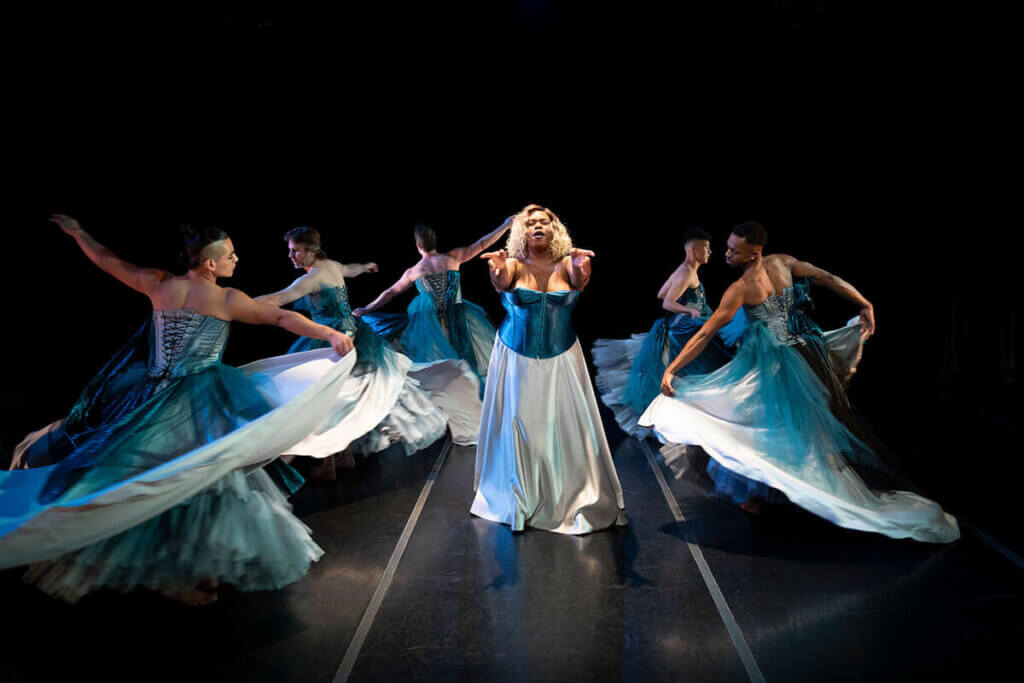 Image is a color photograph of 6 dancers dancing on a black-floored stage. In the center of the photo is a Black trans woman wearing a turquoise corset and floor-length silver skirt, with shoulder-length brown and blond hair, singing and holding her arms out emotionally toward the camera. Surrounding her, 5 queer and trans and gender-nonconforming dancers (Latinx, white, white, Asian-American and Black) spin and spin—all of them are wearing elaborate turquoise, blue, and silver strapless ballgowns. As the 5 dancers spin around, their many layers of skirts twirl up in the air.