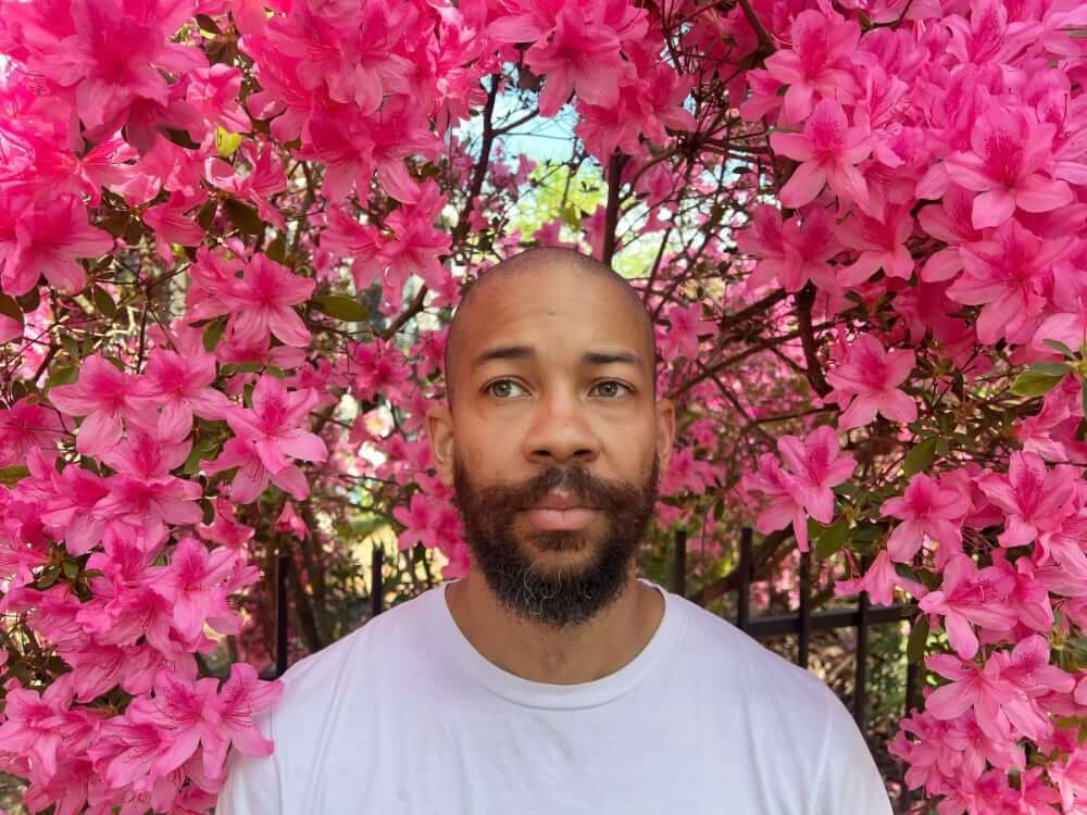 The photo shows a medium-skinned African American man in a white T-shirt. He has a shaven head and dark beard and stands immersed in an azalea bush full of pink blossoms. His stare is pensive as he gazes off into the distance.