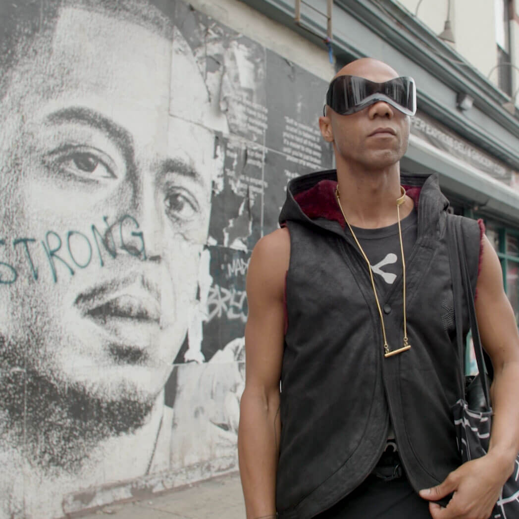 A young Black man with a shaved head and sleeveless, futuristic hooded vest stands on a sidewalk in NYC. His eyes are covered in large, hourglass-shaped wraparound shades, and the lining of the hood is maroon. Behind him is a large black-and-white mural of an unidentified Black man's face, with the word "STRONG" spray painted across the subject's cheek.