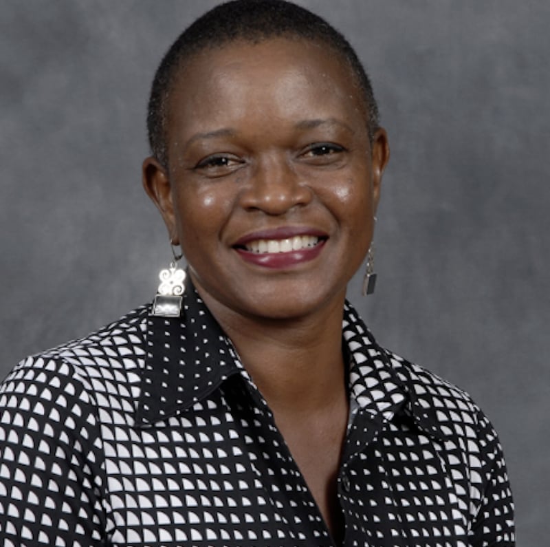 The photo shows a dark-skinned African American woman with short-cropped, natural hair. She is wearing a black-and-white buttoned shirt and silver earrings and smiling broadly at the camera.