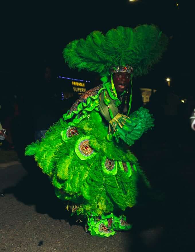 A Masking Indian dances in a dark city street with his arms swinging in a green feathered multicolor rhinestoned suit. His left wing is spread in a swooping curve at the center of the photo. He wears a round feathered sun hat on his head. His eyes are fixed on something to his right, and his mouth is open mid-yell.