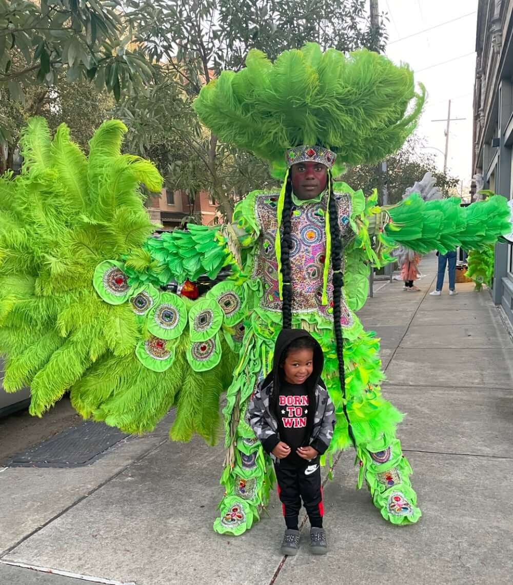 A Masking Indian posing on a sidewalk with his arms wide-open in a green-feathered multicolor rhinestoned suit. His wing is spread on the left side of the photo under his feathered arm and his war spear is on the right side of the photo in his hand. He wears a round feathered sun hat on his head with a multi-green feathered war skirt around his waist. His eyes are fixed on the camera. A small dark-skinned child stands between his legs smiling at the camera in a hoodie and track pants.