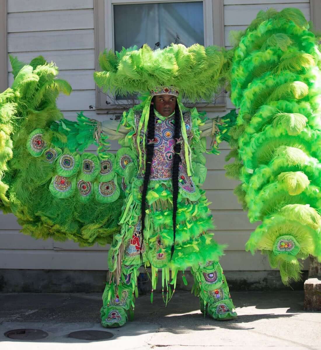 A Masking Indian posing in front of a white paneled home with his arms wide-open in a green-feathered multicolor rhinestoned suit. His wing is spread on the left side of the photo under his feathered arm and his war spear is on the right side of the photo in his hand. He wears a round feathered sun hat on his head with a multi-green feathered war skirt around his waist. His eyes are fixed on his Big Chief awaiting a signal to move out and begin the journey across Midcity.