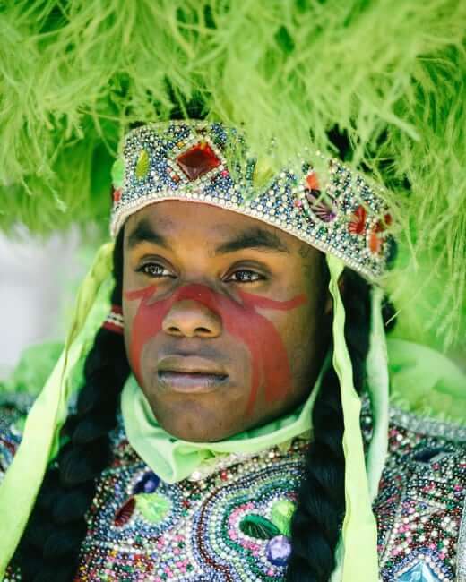 A close-up portrait of a Masking Indian looking left of the camera in a green-feathered multicolor rhinestoned suit. He wears a round feathered sun hat on his head, and the part of the suit covering his chest is covered in rhinestoned patches. Two decorated braids run down his chest and out of the frame.