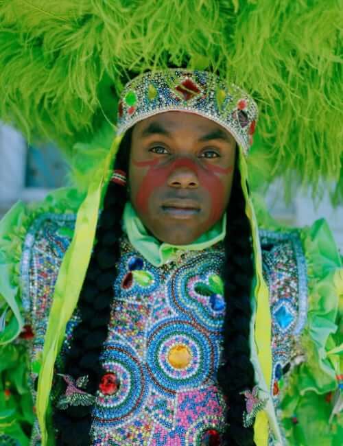 A close-up portrait of a Masking Indian looking directly at the camera in a green-feathered multicolor rhinestoned suit. He wears a round feathered sun hat on his head, and the part of the suit covering his chest is covered in rhinestoned patches. Two braids decorated with hummingbird pins run down his chest and out of the frame.