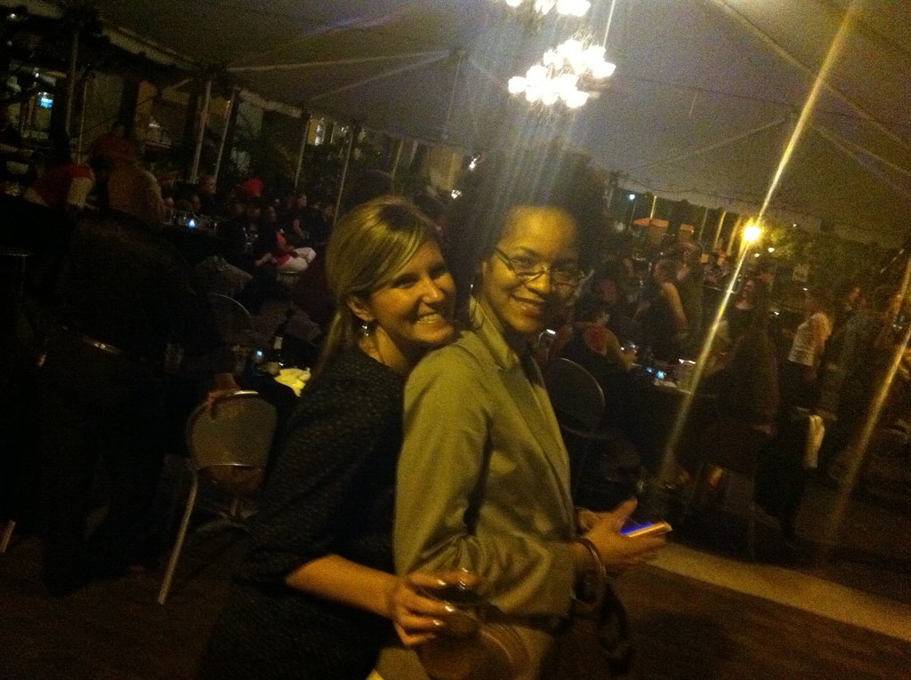 Stephanie Atkins, a Black woman, poses in profile with a white woman behind her who is looking over Stephanie's shoulder at the camera. Both women are smiling. Stephanie is holding a smartphone, and the other woman is holding a drink. It is nighttime, and behind them is a temporary tent structure with several dozen people gathered in a celebration.