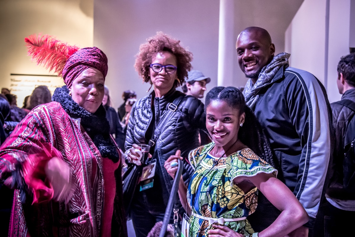 A group of four Black people pose for a photo at an indoor event. On the left, a woman wears a red patterned wrap, a black scarf, and a matching headwrap adorned with a large red feather. Next to her is Stephanie Atkins, wearing glasses and a black quilted jacket. To her right, a younger woman with dark, braided hair wears a colorful, patterned dress. Behind and to the right of her, a man with a shaved head and a gray scarf smiles toward the camera. Other people are partially visible in the background, which appears to be a gallery or event space with white walls.