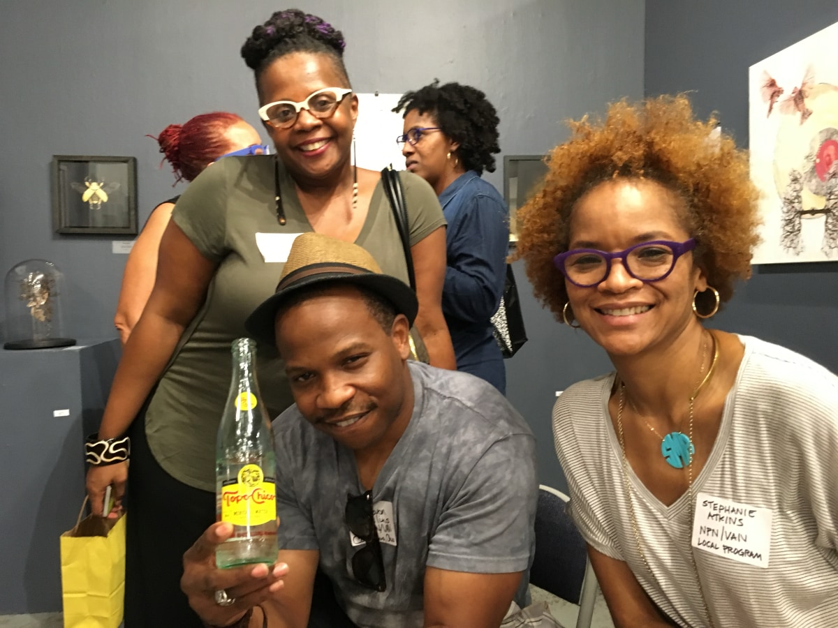 Stephanie Atkins, a Black woman, poses with a Black man and another Black women in an art gallery. Stephanie leans in from the right. In the lower middle of the photo, the Black man, who is wearing a tan and black fedora, leans forward and smiles as he holds up a bottle of Tope Chico mineral water. The other Black woman stands behind him with a small yellow paper shopping bag in her right hand. Two other women of color stand in the background, partially obscured by the trio.