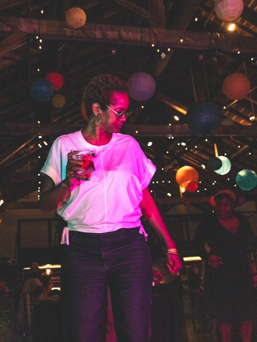Stephanie Atkins, a Black woman wearing a white blouse and dark pants, stands in an indoor space that has been decorated for a celebration, with round paper lanterns of various colors hanging above her, and strings of clear lights wrapped around the rafters. The space is fairly dark, and Stephanie is illuminated by a bright pink light. She is holding a drink in one hand and looking off to the side.