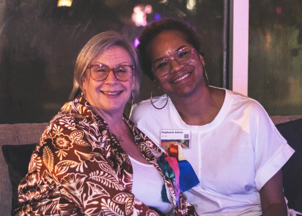 Stephanie Atkins, a Black woman, sits next to a white woman on a couch at what appears to be a nighttime event. The two women are leaning into each other and smiling warmly at the camera.
