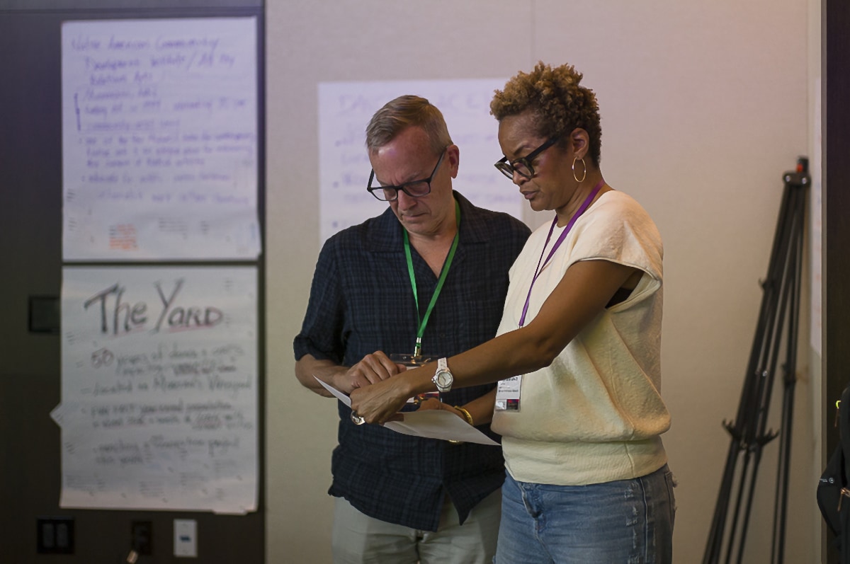 Stephanie Atkins, a Black woman wearing a short sleeved white blouse and denim blue jeans, stands next to a white man wearing a blue button-down shirt and gray pants. They are in what appears to be a conference room. Stephanie is holding a piece of paper in front of them and they are both staring at it. Behind them, affixed to the wall, are large pieces of white paper covered in writing.